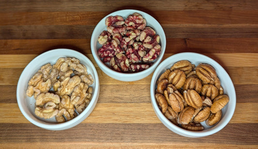 Three small white bowls on a wooden surface, each containing different types of nuts: one with walnuts, one with pecans, and one with red walnuts.