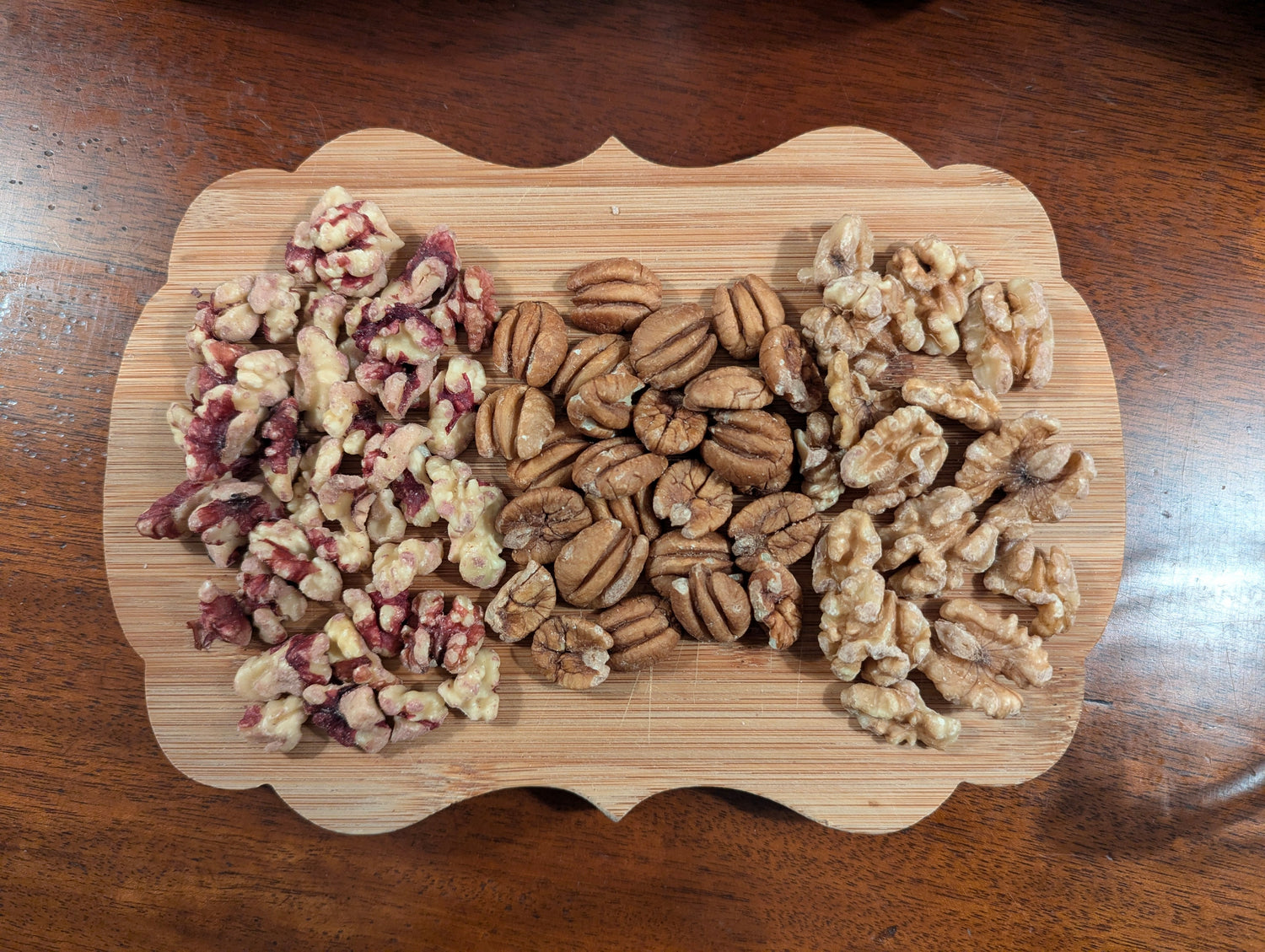 A wooden board with three piles of nuts: chopped pecans on the left, whole pecans in the center, and walnut halves on the right. The board is on a wooden surface.