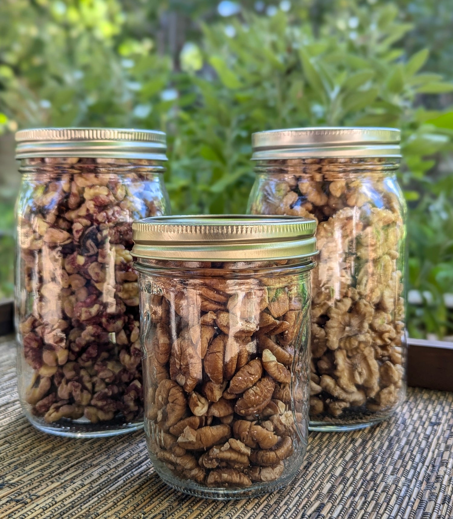 Three glass jars filled with different types of nuts are placed on a woven surface. A green, leafy background can be seen behind them.