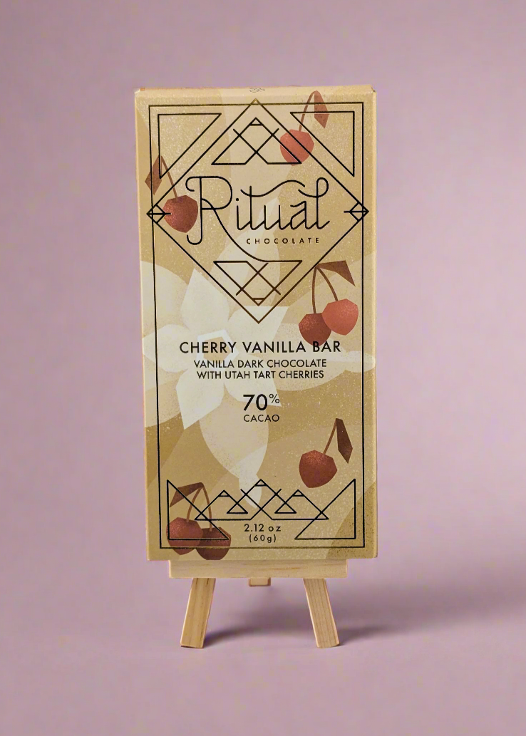 A bar of Ritual Chocolates Cherry Vanilla Bar with a decorative geometric gold and cherry design, displayed on a small wooden easel against a soft purple background. The package notes its 70% cacao, 2.12 oz.