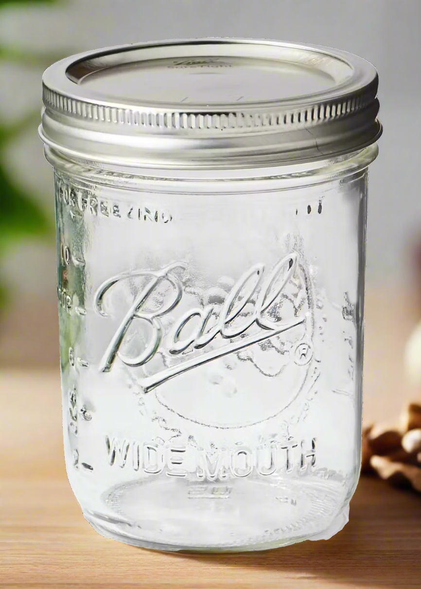 A Ball brand Mason jar with Wide Mouth embossing sits on a wooden surface, its clear glass and metal lid pairing elegantly. Blurred green leaves and a nut are visible in the backdrop.