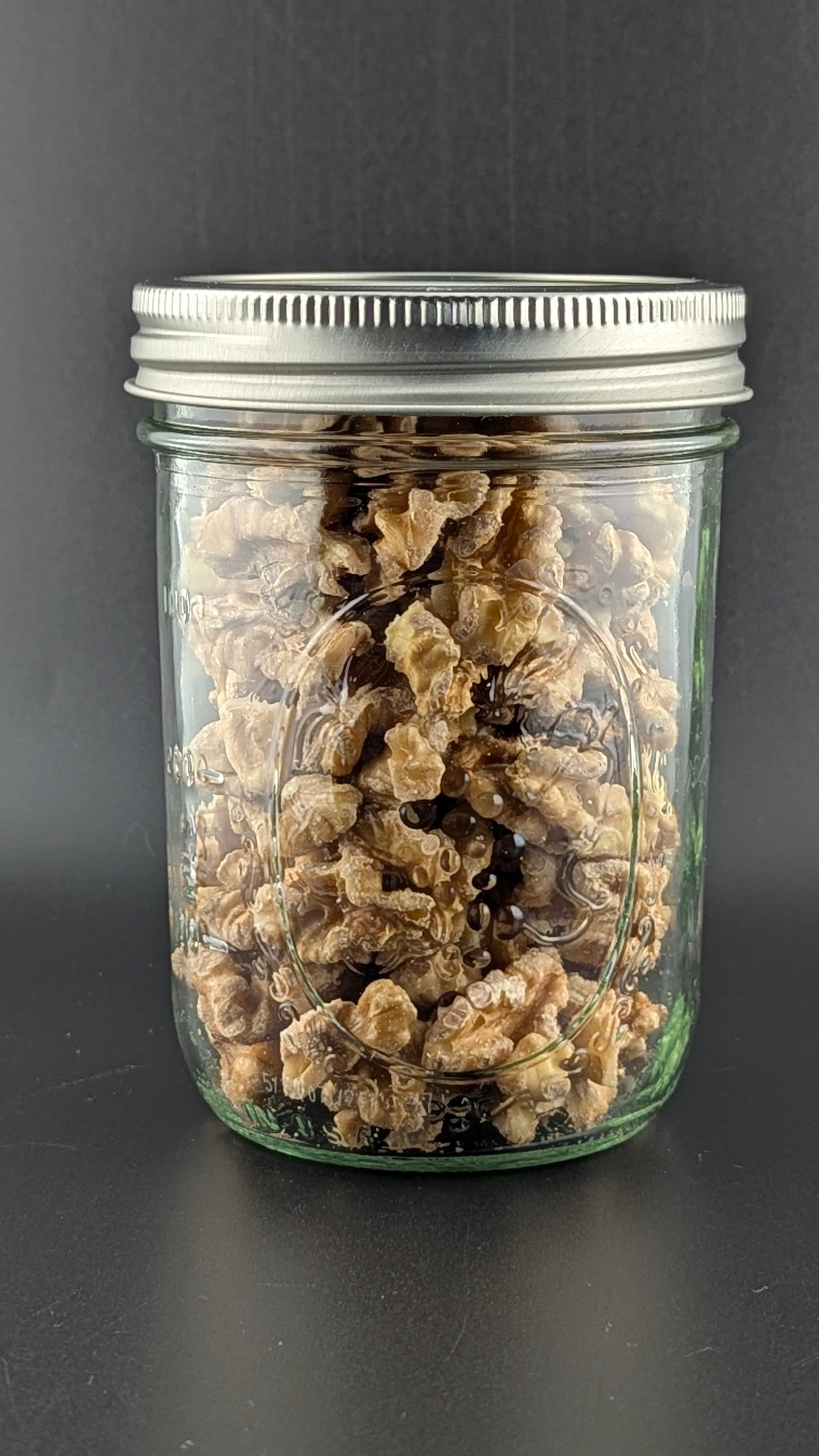 A clear glass jar filled with dried mushrooms sits against a dark background under its gleaming silver lid. The densely packed mushrooms resemble the rich, textured look of organic Infused Chandler Walnuts.