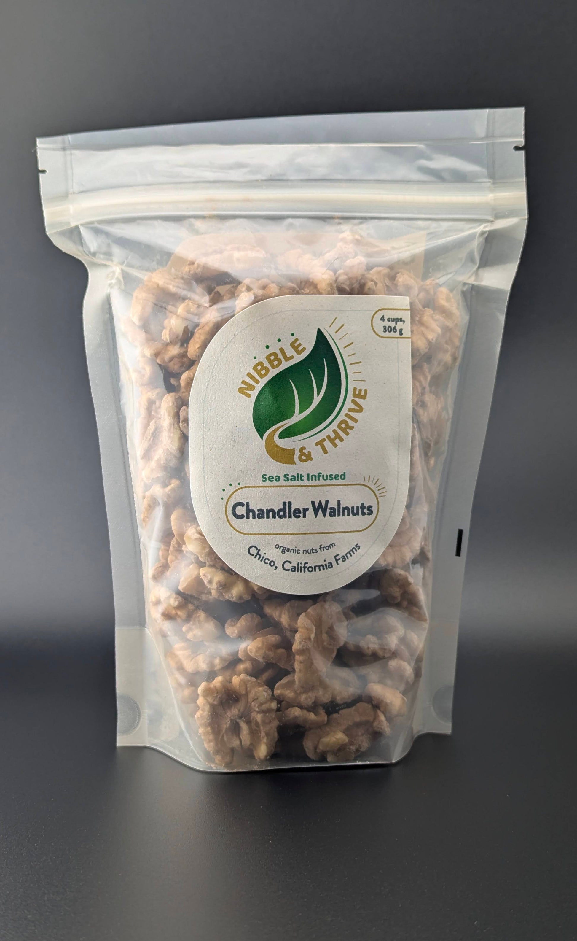 A clear bag of Nibble & Thrives Infused Chandler Walnuts, Organic with a green leaf label is showcased on a dark background. These omega-3-rich, sea salt-infused walnuts are from Chico, California.