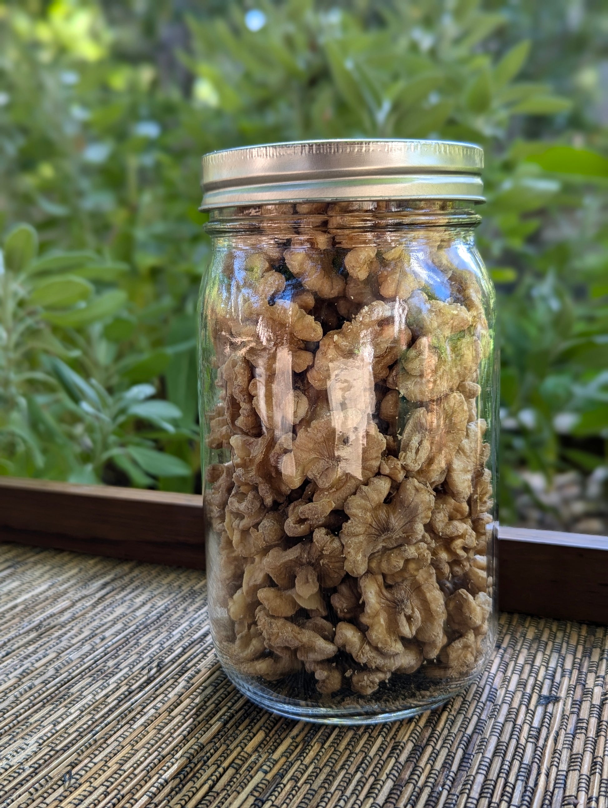 A clear glass jar of Organic Infused Chandler Walnuts with a metal lid rests on a woven mat. The natural light highlights the wholesome nuts against lush green foliage, promising a rich dose of omega-3s.