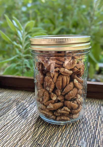 A Mason jar (Ball) filled with pecans rests on a woven mat. The jar, with a gold lid, is set outdoors against lush green foliage, creating a serene and natural vibe.