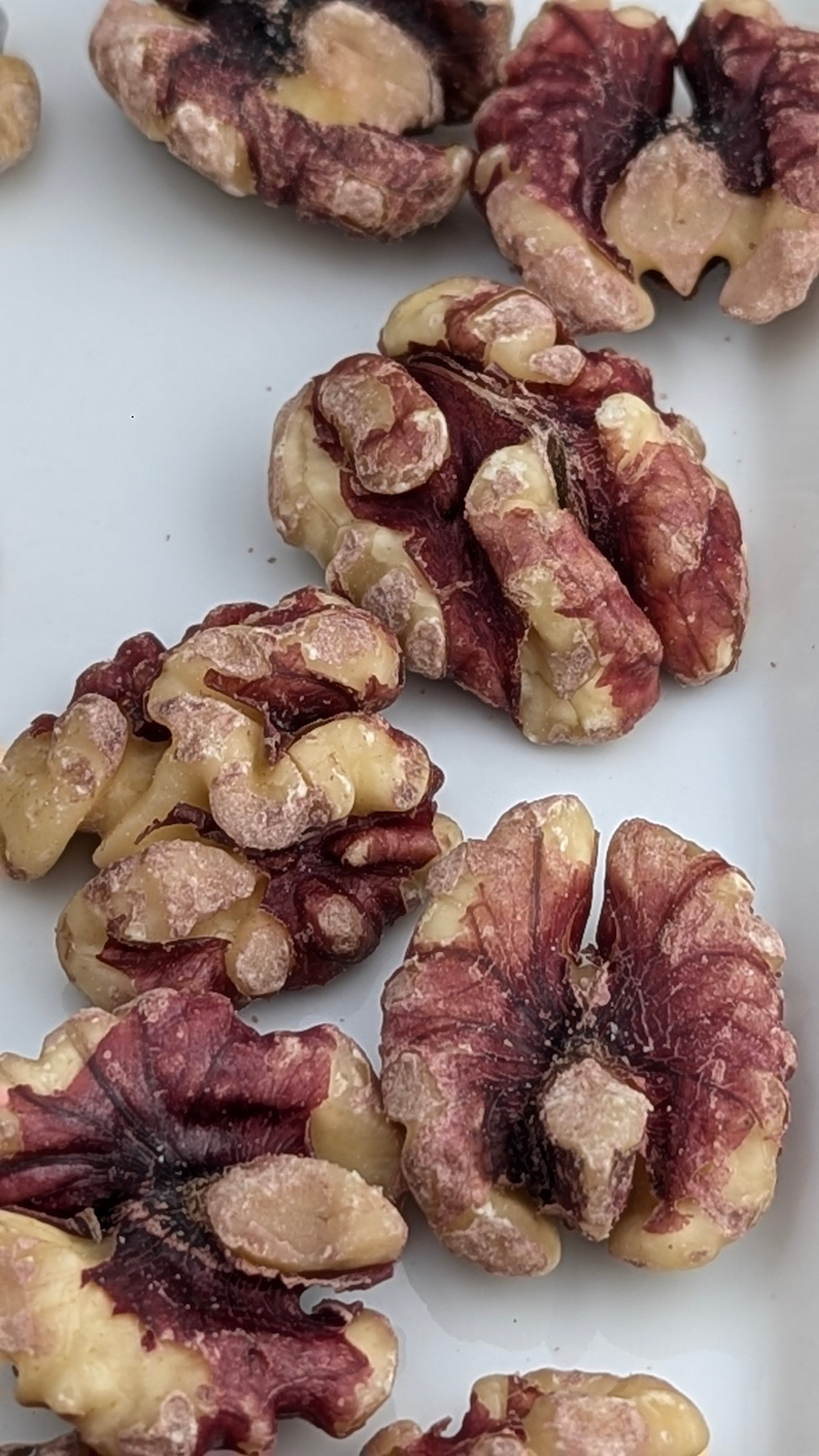 Close-up of several Infused Red Walnuts, Organic, scattered on a white surface. These walnuts have a unique deep red skin and light brown inner flesh, making them the perfect nutritious snack for any occasion.