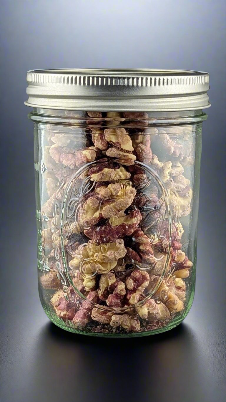 The jar of Infused Red Walnuts, Organic, features a metal lid and is filled to the brim with shelled walnuts. It sits on a smooth gray surface, allowing the nutritious walnuts to be clearly visible through the transparent glass.