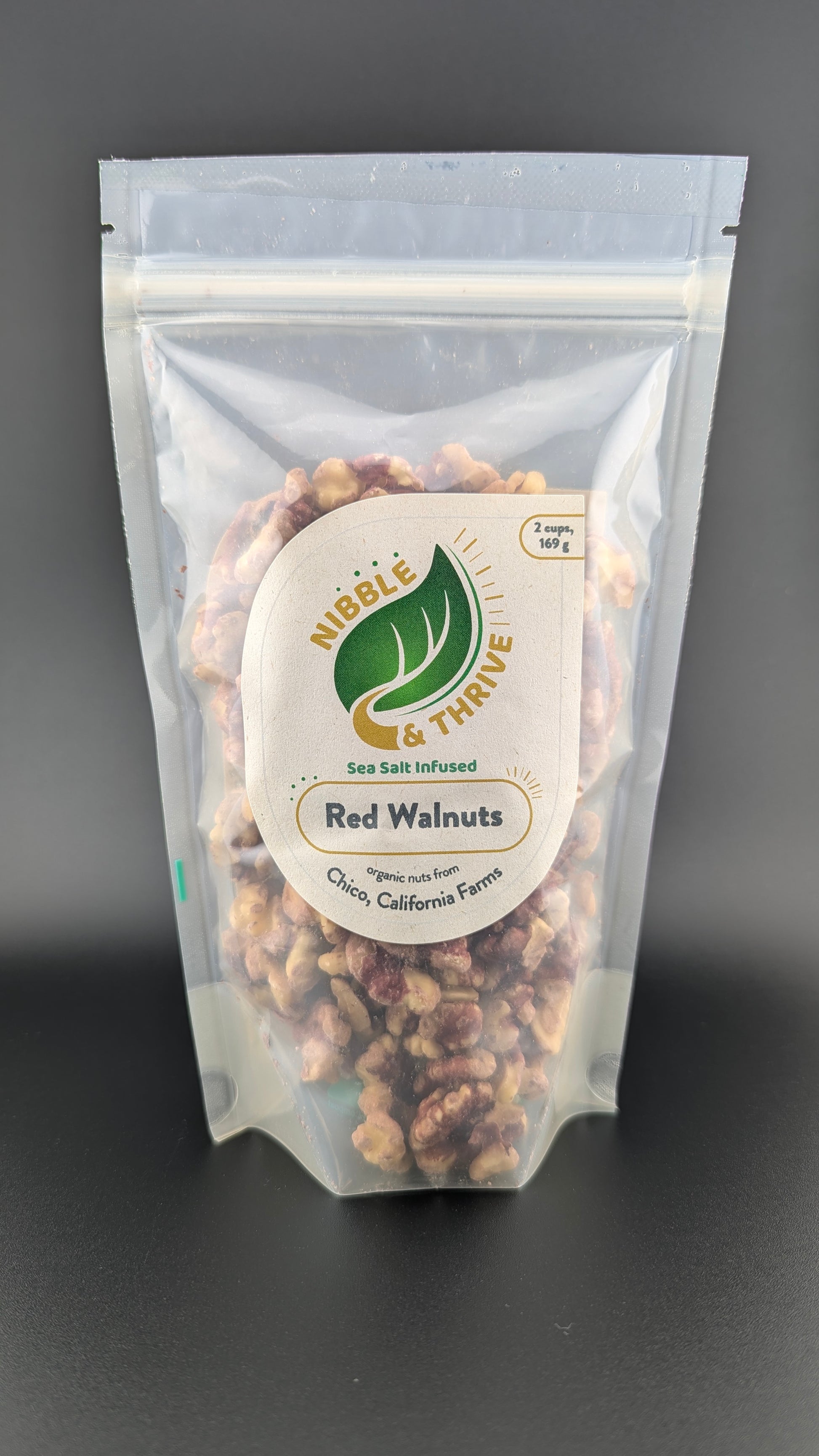 A clear, resealable Nibble & Thrive bag holds organic sea salt-infused red walnuts from Chico, California Farms. The vibrant nuts are showcased against a dark background.