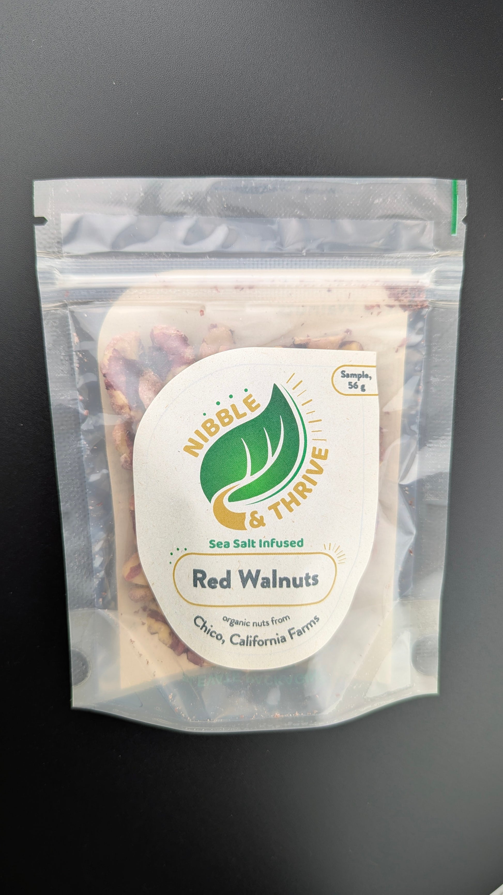 A sealed 56g pack of Nibble & Thrives Infused Red Walnuts, Organic with sea salt features a green leaf logo and organic nuts from Chico, California Farm label. Against a dark background, this tasty snack is highlighted in our walnut clearance sale.