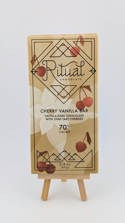 A Ritual Cherry Vanilla Bar 70%, 2.12 oz sits on a small easel, featuring cherry illustrations and text stating vanilla dark chocolate blends with Utah Montmorency cherries.