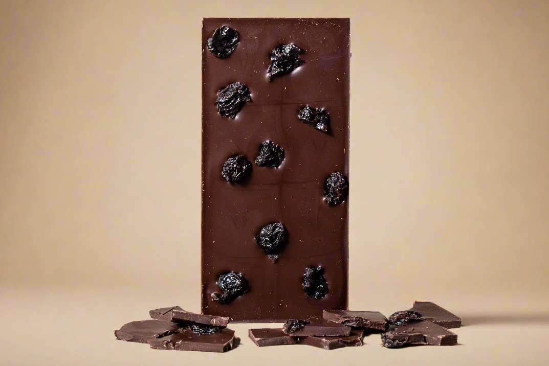 A Ritual Cherry Vanilla Bar 70%, 2.12oz (60 g) stands on a light beige surface, featuring dark nuances and Montmorency cherries. Scattered at the base are small pieces of chocolate, highlighting the exquisite blend.