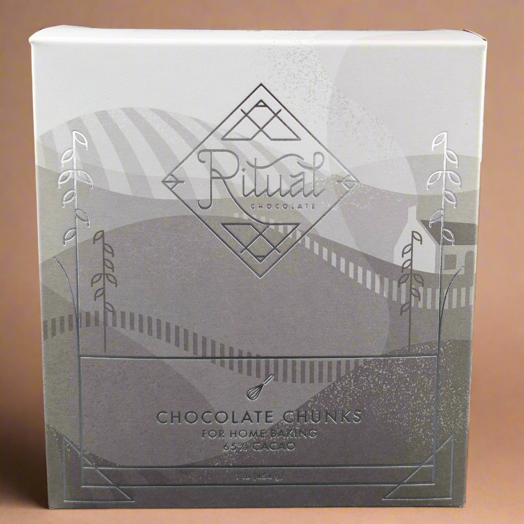 A 1 lb box of Ritual Chocolate Chunks - Chefs Blend 65% is crafted for home baking with organic cacao beans, featuring elegant geometric packaging and abstract illustrations of fields and hills.