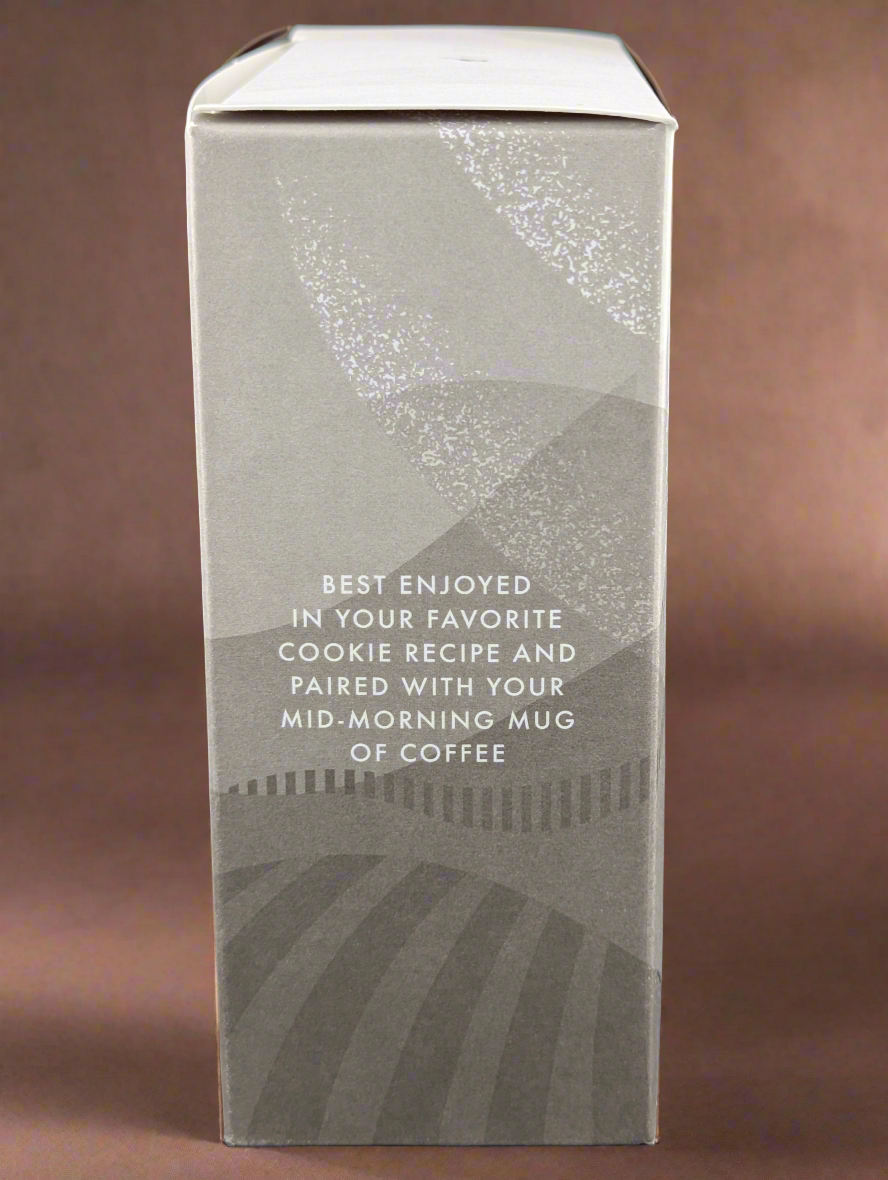 A gray-themed side view of the Ritual Chocolate Chunks - Chefs Blend 65% box shows abstract designs. The text suggests enjoying it in your cookie recipes alongside a mid-morning coffee. The background is a soft brown surface.