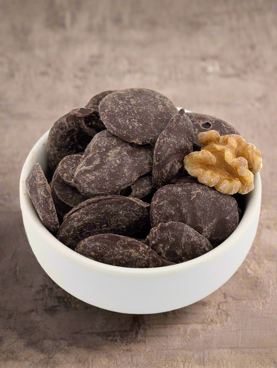 A white bowl with Ritual Chocolate Chunks - Chefs Blend 65% sits on a textured surface, filled with dark chocolate chunks made from organic cacao beans. A walnut half rests atop the pieces, creating a contrasting scene.