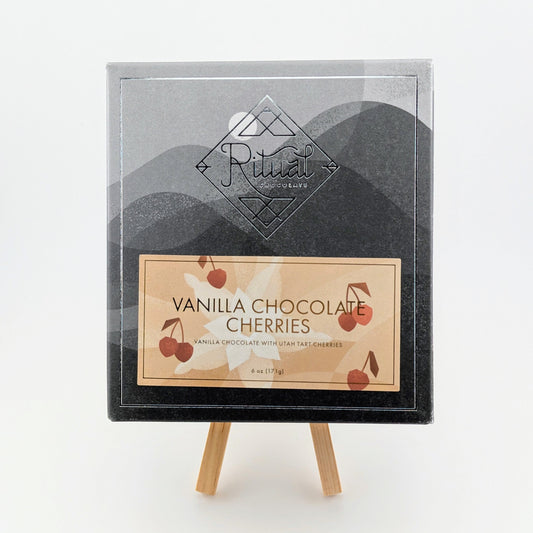 A box of Ritual Vanilla Chocolate Cherries - 6 oz (171 g) sits on a small easel. The packaging showcases a geometric design featuring Utah tart cherries and mountains, all delicately wrapped in vanilla.